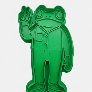 May include: A green frog-shaped cookie cutter. The frog is wearing a button-down shirt and pants, and is giving a peace sign with its hand. The cookie cutter is made of plastic and is designed to cut out the shape of a frog.