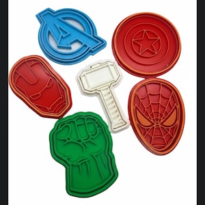 May include: A collection of colorful cookie cutters featuring superhero emblems. Includes the Avengers "A" logo in blue, Captain America's shield in red, Iron Man's mask in red, Thor's hammer in white, the Hulk's fist in green, and Spider-Man's mask in red.