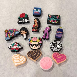 May include: A collection of colorful rubber shoe charms featuring various designs including a rainbow with text, a rose, a portrait with flowers, heart-shaped bread patterns, a purple outfit figure, and rectangular plaques with Spanish text.