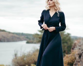 May include: A woman wearing a long, dark blue wrap dress with long sleeves. She is standing on a rocky surface with a body of water in the background.