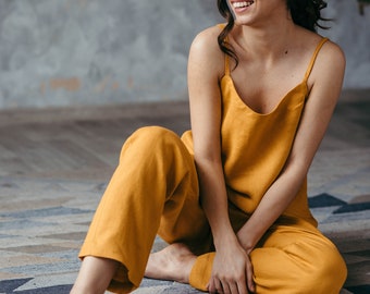 Puede incluir: Una mujer con un mono amarillo mostaza se sienta en una alfombra con estampado con las piernas cruzadas. Sonríe y mira hacia un lado.