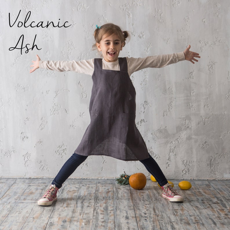 A child wearing a grey linen pinafore apron, looking playful in a well-lit studio space.