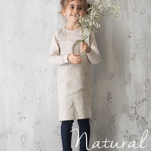 A young child wearing a natural linen apron, standing happily in a neutral studio.
