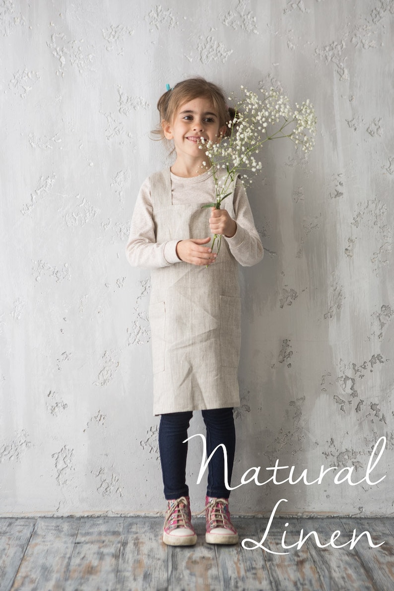 A young child wearing a natural linen apron, standing happily in a neutral studio.
