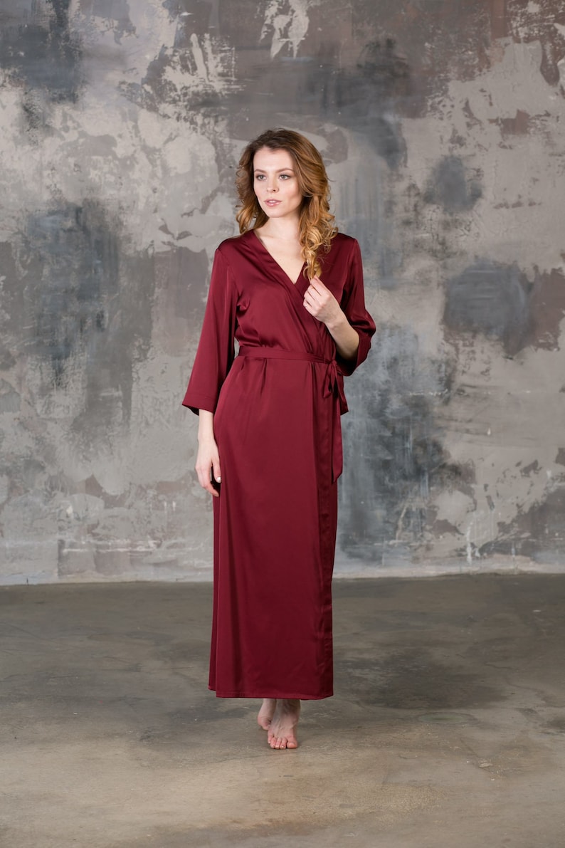 May include: A long, burgundy satin robe with a wrap closure and long sleeves. The robe is worn by a person standing in front of a textured gray wall.