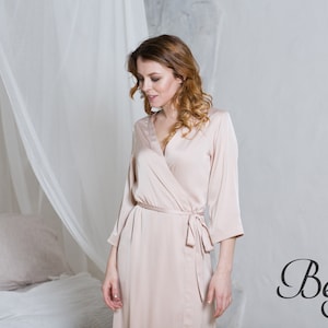 May include: A beige satin robe with a long sleeve and a tie belt. The robe is worn by a person standing in a bedroom.