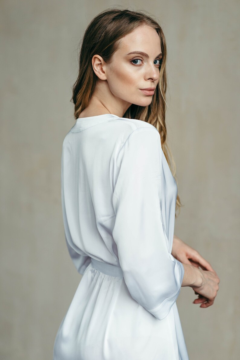 May include: A woman wearing a white satin robe with a sash tied at the waist. The robe has long sleeves and a V-neckline.