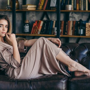 May include: A woman wearing a light brown satin robe is sitting on a brown leather couch. She is looking at the camera with a thoughtful expression.