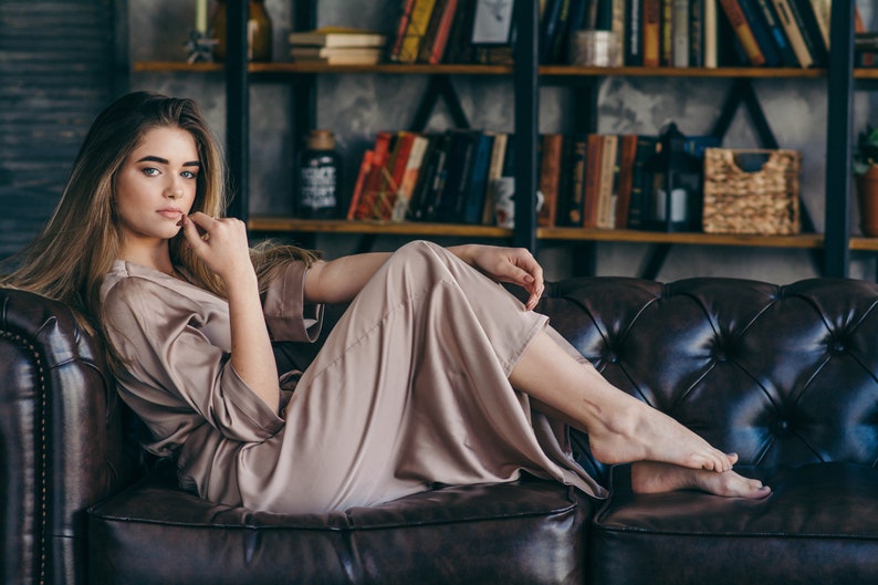 May include: A woman wearing a light brown satin robe is sitting on a brown leather couch. She is looking at the camera with a thoughtful expression.