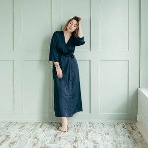 May include: A woman wearing a long, dark blue satin robe with a tie belt. She is standing in a room with white walls and a window with white blinds.