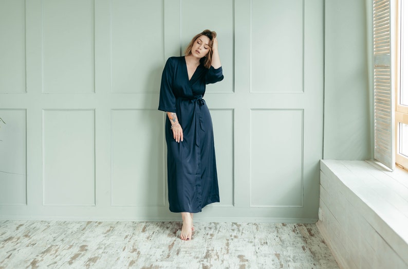 May include: A woman wearing a long, dark blue satin robe with a tie belt. She is standing in a room with white walls and a window with white blinds.