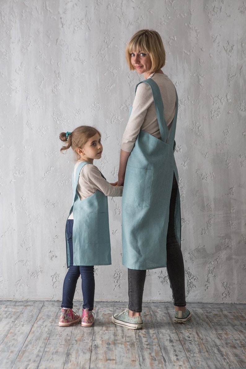 An adult and child in matching grey linen pinafore aprons, highlighting the cross-back design in a clean, neutral space.