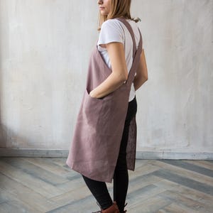 May include: A dusty rose linen apron with large front pockets and long straps that cross in the back. The apron is worn over a white t-shirt and black pants. The model is wearing brown boots.