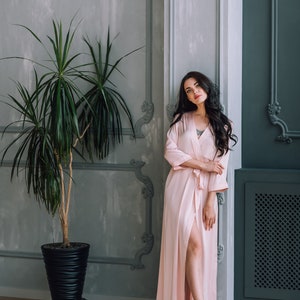 May include: A woman in a light pink satin robe stands in a room with a potted plant and gray and blue walls. She is looking at the camera.
