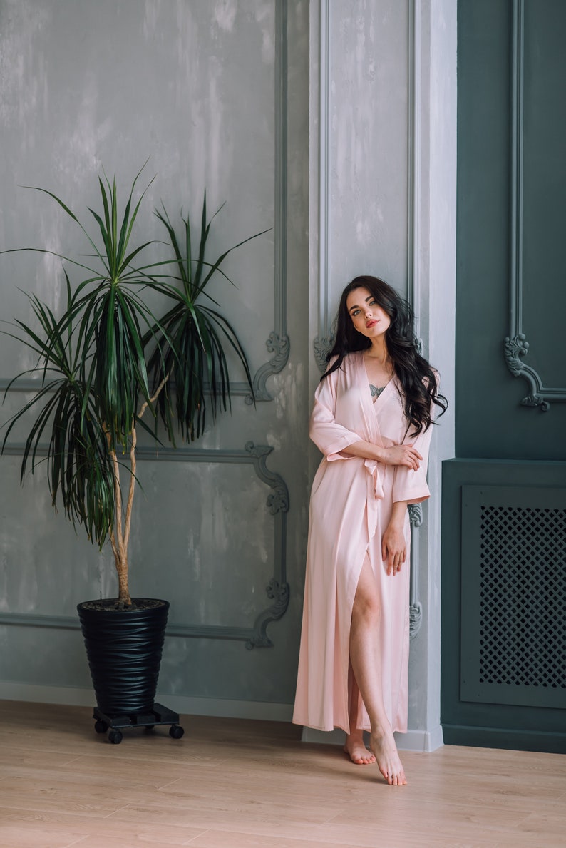 May include: A woman in a light pink satin robe stands in a room with a potted plant and gray and blue walls. She is looking at the camera.