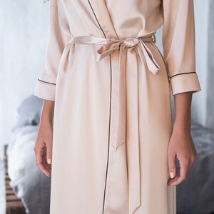 May include: A long, light beige satin robe with a self-tie sash and black piping around the edges.