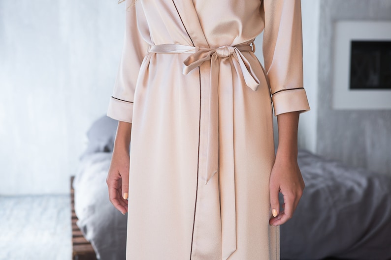 May include: A long, light beige satin robe with a self-tie sash and black piping around the edges.