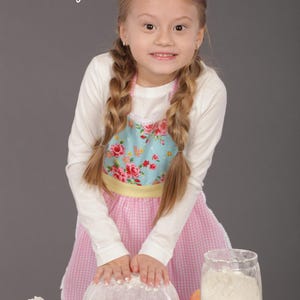 Little girl smiling and holding bowl wearing pink apron with yellow trim