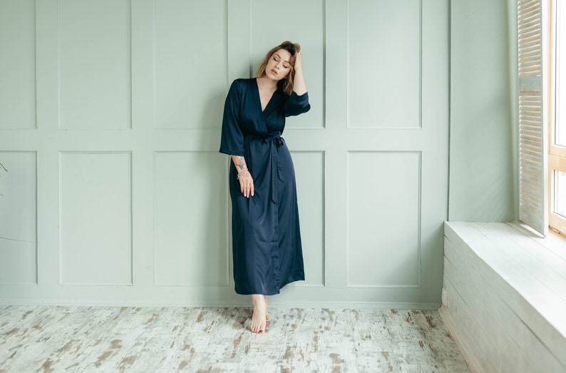 May include: A woman wearing a long, dark blue satin robe with a sash tie. She is standing in front of a light green wall with white trim.