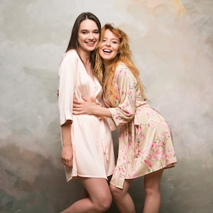 Op de afbeelding: Twee vrouwen dragen bijpassende badjassen, een in een effen roze badjas en de andere in een badjas met bloemenprint, ze glimlachen en omhelzen elkaar. Ze staan voor een textuur achtergrond.