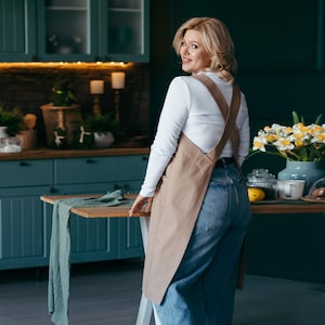 Pode incluir: Uma mulher vestindo um avental marrom com uma blusa branca e jeans azul está em uma cozinha. Ela está olhando por cima do ombro para a câmera. Há uma mesa com um pano verde na frente dela.