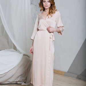 May include: A woman wearing a long, light pink satin robe with a tie belt. She is standing on a round, woven rug in a bedroom.