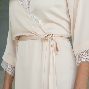 May include: A soft, cream-colored satin robe with delicate lace trim on the sleeves and neckline. The robe has a tie belt and a relaxed fit.