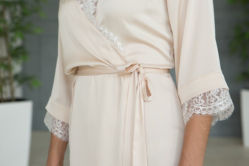 May include: A soft, cream-colored satin robe with delicate lace trim on the sleeves and neckline. The robe has a tie belt and a relaxed fit.