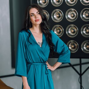 May include: A woman wearing a teal blue satin robe with a long sleeve and a tie belt. She is standing in front of a wall with a grid of round silver accents.