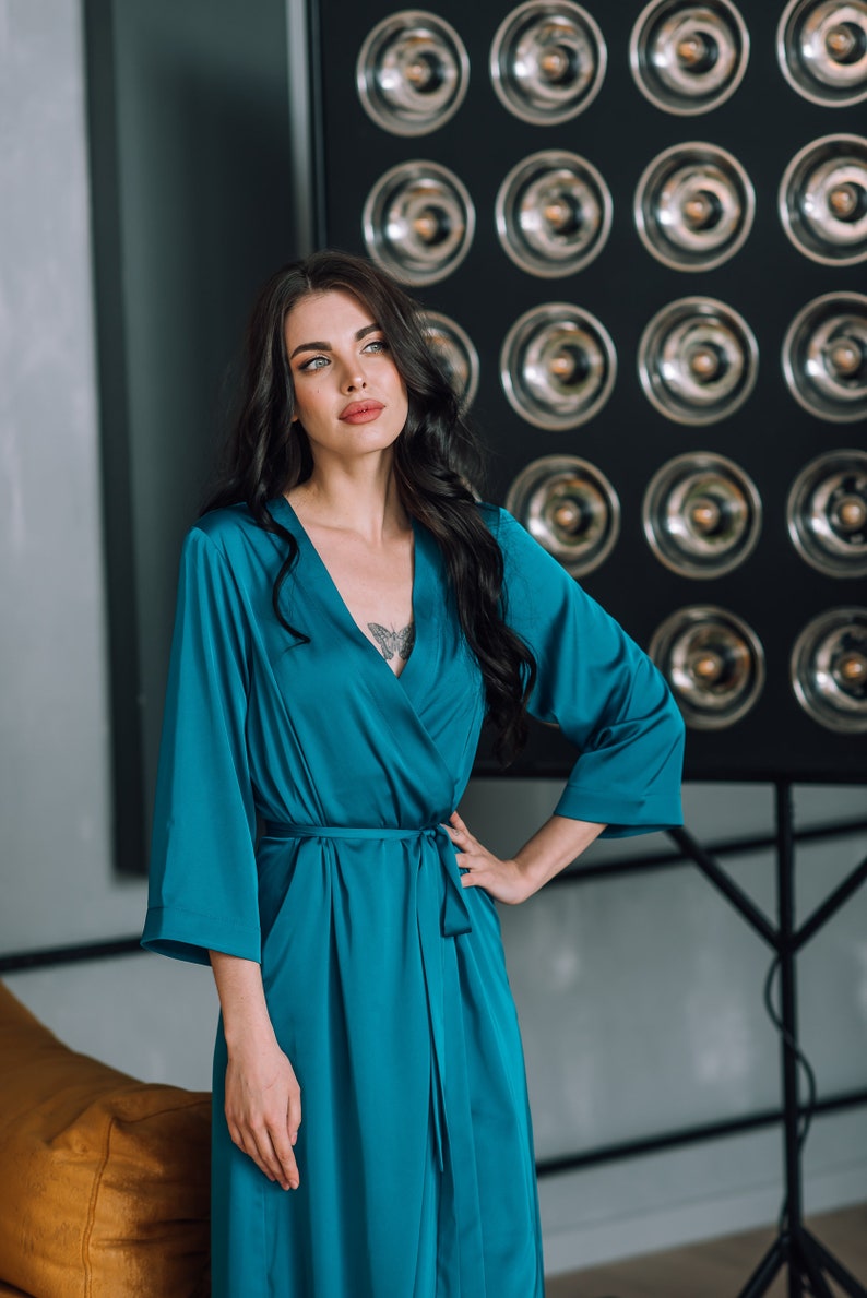 May include: A woman wearing a teal blue satin robe with a long sleeve and a tie belt. She is standing in front of a wall with a grid of round silver accents.