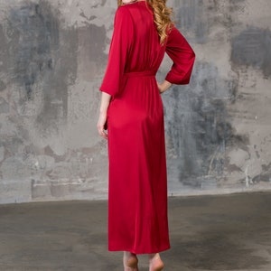 May include: A long, red satin robe with a tie belt. The robe is worn by a person standing in front of a gray concrete wall.