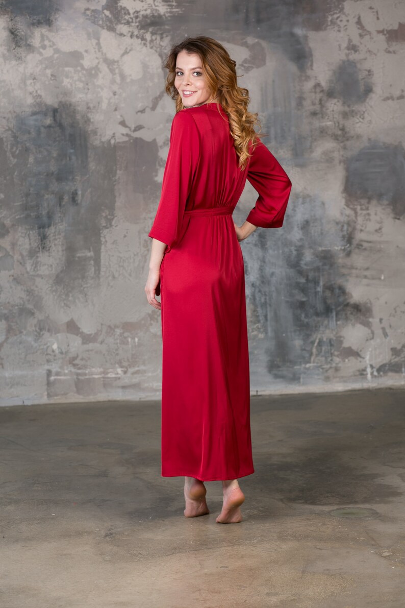 May include: A long, red satin robe with a tie belt. The robe is worn by a person standing in front of a gray concrete wall.