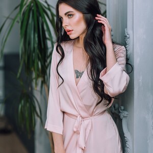 May include: A woman wearing a light pink satin robe with a butterfly tattoo on her chest. The robe has a tie belt and long sleeves.