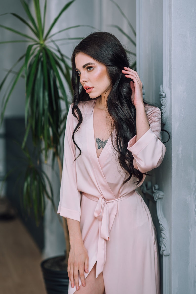 May include: A woman wearing a light pink satin robe with a butterfly tattoo on her chest. The robe has a tie belt and long sleeves.
