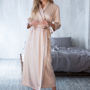 May include: A long, light beige satin robe with a black trim. The robe has a shawl collar and a tie belt. The model is standing on a wooden floor.