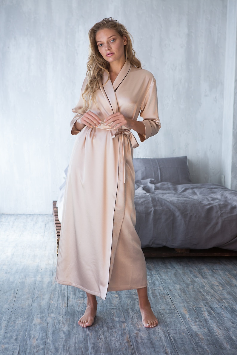 May include: A long, light beige satin robe with a black trim. The robe has a shawl collar and a tie belt. The model is standing on a wooden floor.