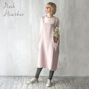 A model in a soft pink cross-back apron, standing in a neutral, well-lit studio.