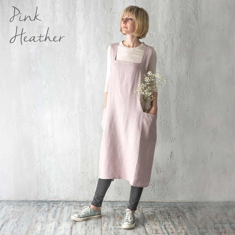 A model in a soft pink cross-back apron, standing in a neutral, well-lit studio.
