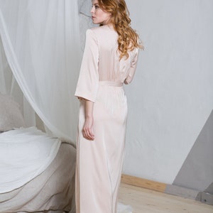 May include: A woman wearing a long, pale pink satin robe with a sash. She is standing on a round, woven rug in a room with white walls and a bed in the background.