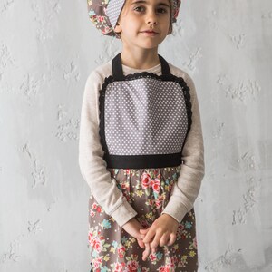 Girl in floral print apron holding whisk and wearing chef hat, family gift idea.