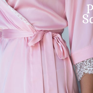 May include: A pink satin robe with white lace trim and a matching sash. The robe is tied at the waist and has long sleeves.