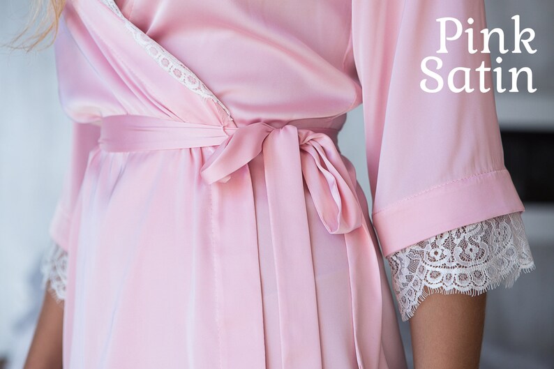 May include: A pink satin robe with white lace trim and a matching sash. The robe is tied at the waist and has long sleeves.