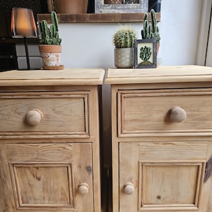 May include: Two matching light-toned wooden bedside cabinets, each featuring a single top drawer and a lower cupboard door. Both units are constructed with visible wood grain, square recessed panels on the doors, and simple round wooden knobs.