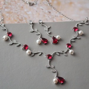 以下が含まれることがあります： つる模様のシルバーネックレスとイヤリングのセット。ネックレスとイヤリングには、赤い宝石と白い真珠のアクセントが施されています。