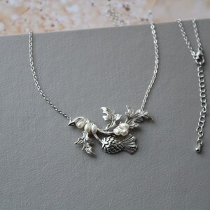 May include: A silver necklace with a thistle pendant and pearl accents. The pendant features a detailed design of a thistle flower with leaves and stems. The necklace is on a silver chain with a lobster clasp.