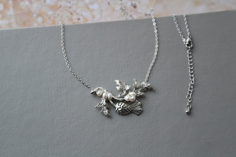 May include: A silver necklace with a thistle pendant and pearl accents. The pendant features a detailed design of a thistle flower with leaves and stems. The necklace is on a silver chain with a lobster clasp.