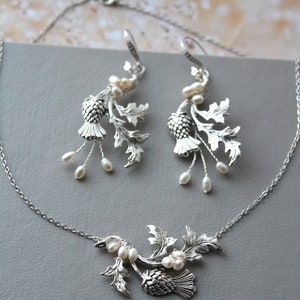 May include: A silver necklace and earrings set featuring a thistle design with white pearl accents. The thistle is a traditional Scottish symbol.