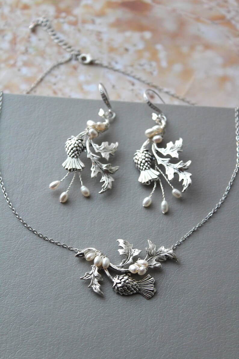 May include: A silver necklace and earrings set featuring a thistle design with white pearl accents. The thistle is a traditional Scottish symbol.