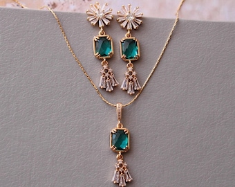 May include: A gold necklace and earrings set with emerald green square-shaped stones and clear crystal accents. The pendant and earrings feature a tassel design with clear crystals.