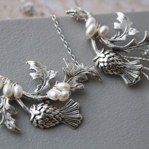 May include: Silver necklace with two thistle pendants and pearl accents. The thistle is a traditional Scottish symbol.